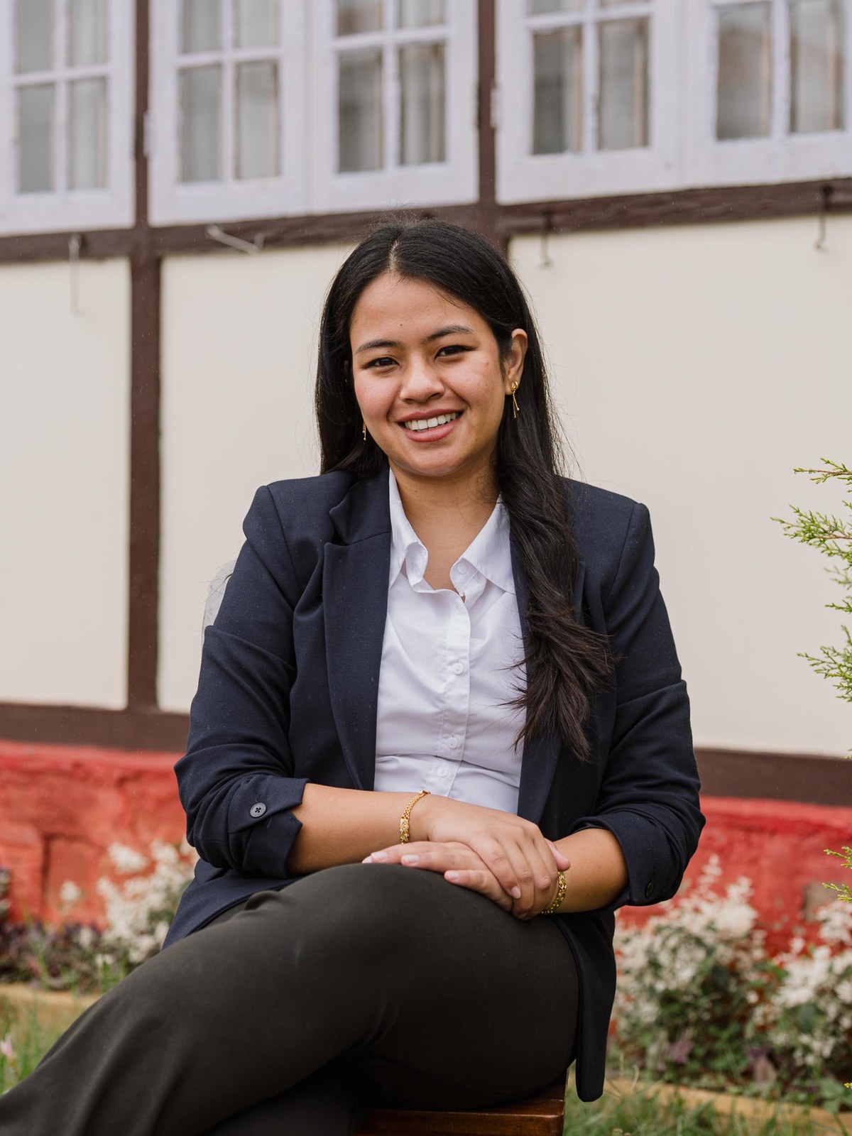 Dr. Emidaka, founder of Iyka-Aram, seated in the clinic garden in a navy blazer and white shirt, the veranda's red plinth and hedge behind her, smiling towards the camera