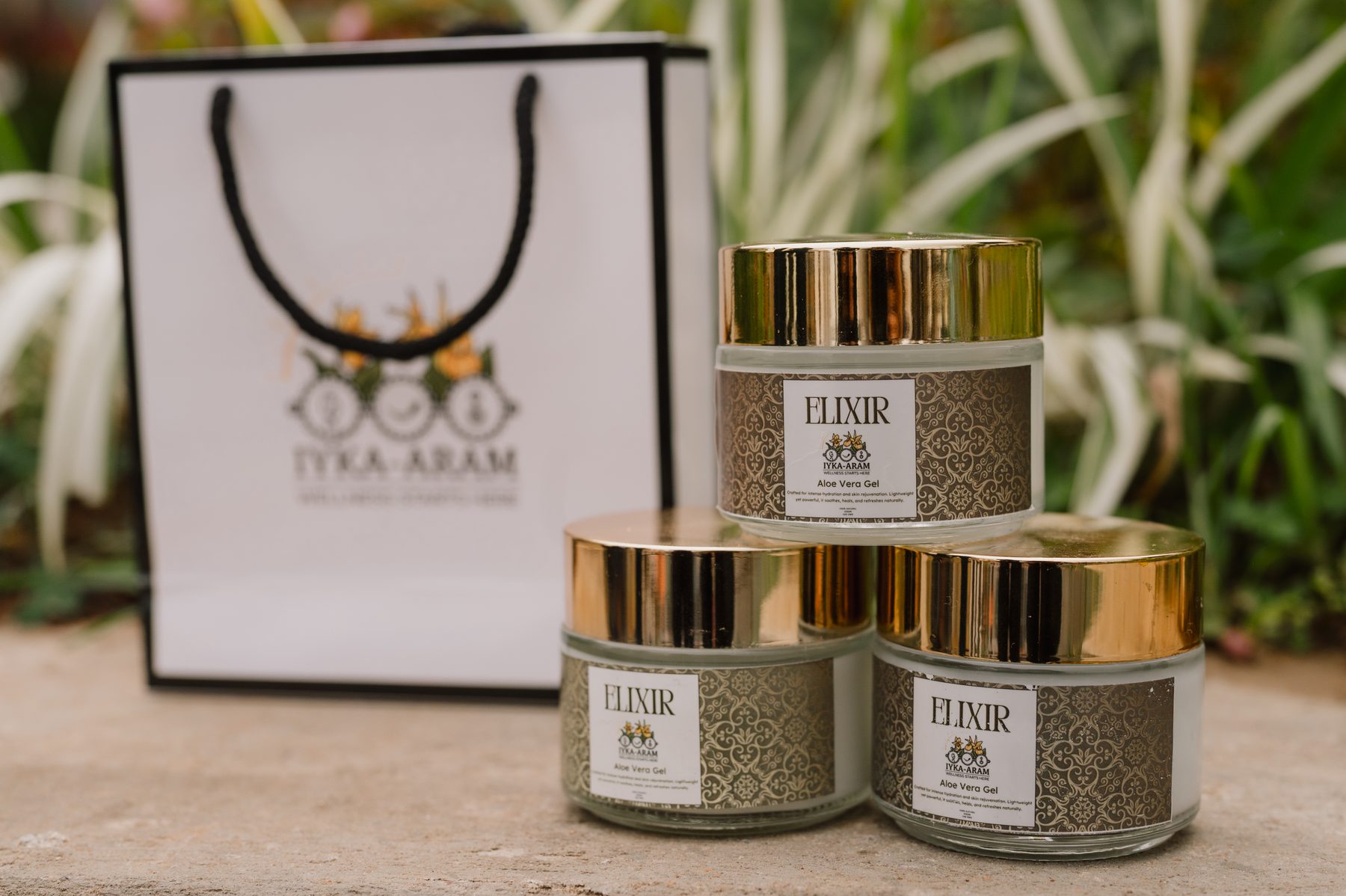 Three gold-lidded jars of ELIXIR Aloe Vera Gel stacked on a stone ledge in front of an Iyka-Aram gift bag, with garden foliage behind