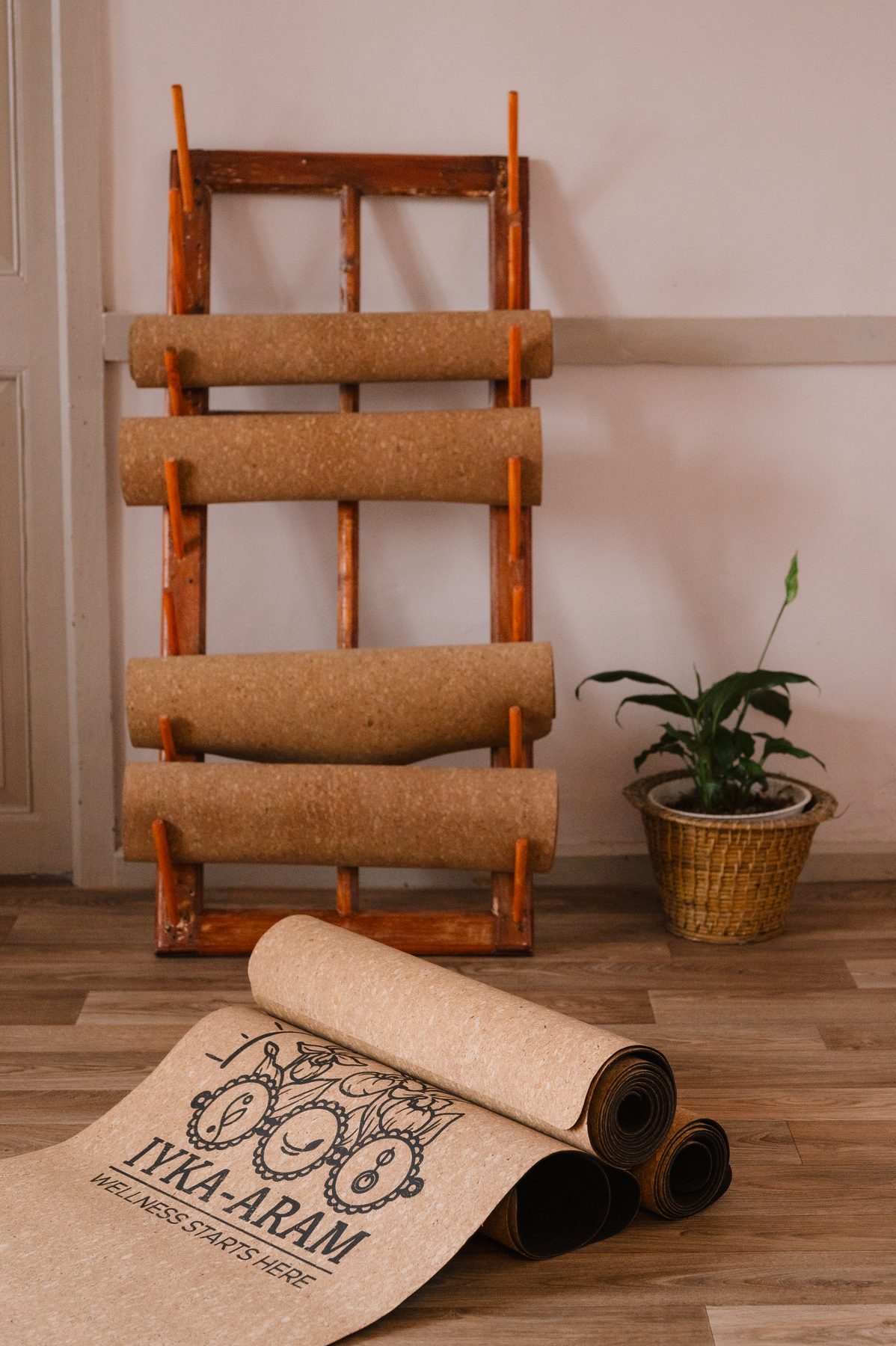 A wooden rack holding rolled cork yoga mats beside a potted plant, with one Iyka-Aram mat unrolled across the wooden floor