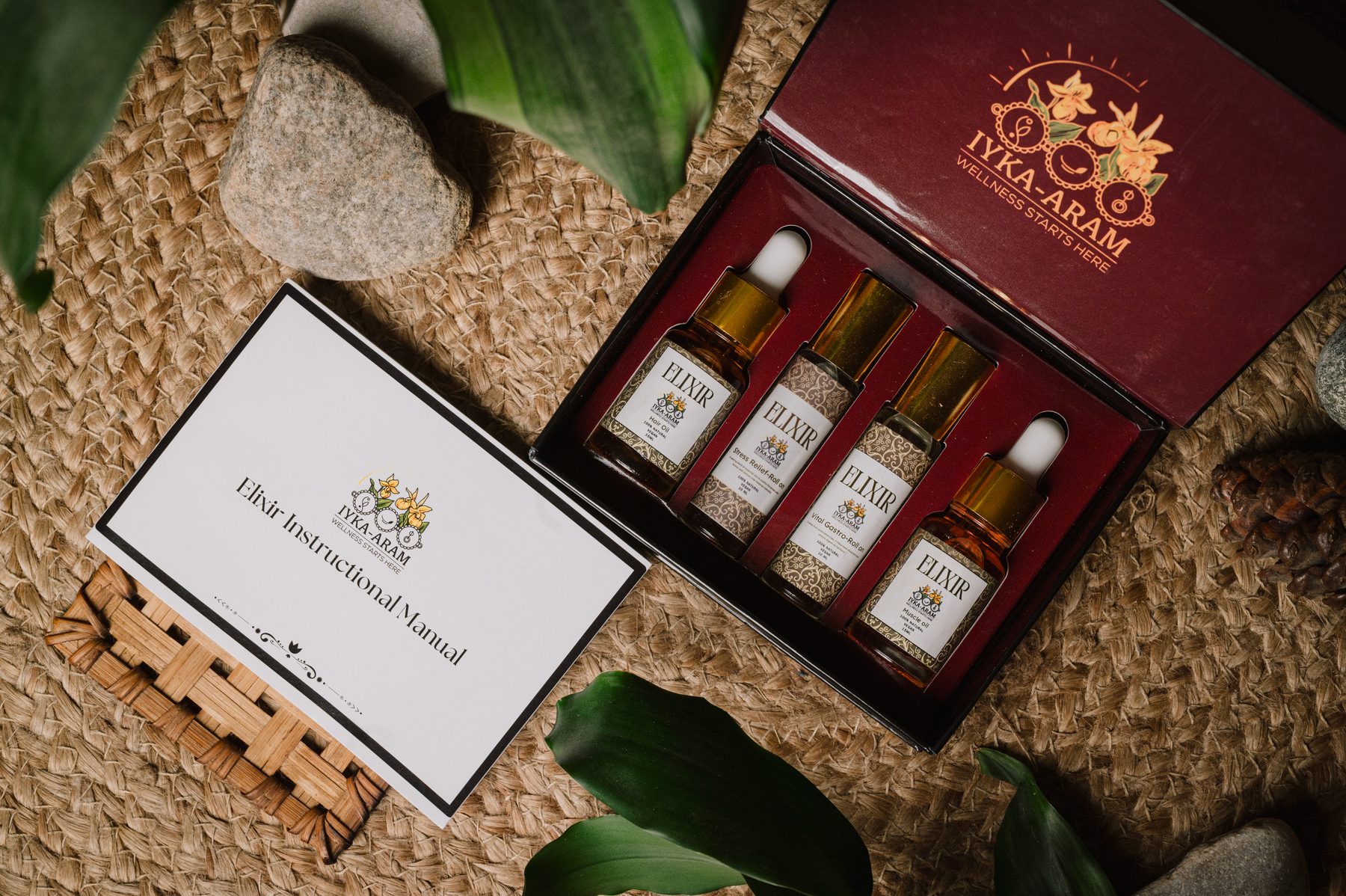 An open crimson-lined Iyka-Aram gift box holding four ELIXIR bottles, photographed from above on woven jute beside the printed Elixir Instructional Manual