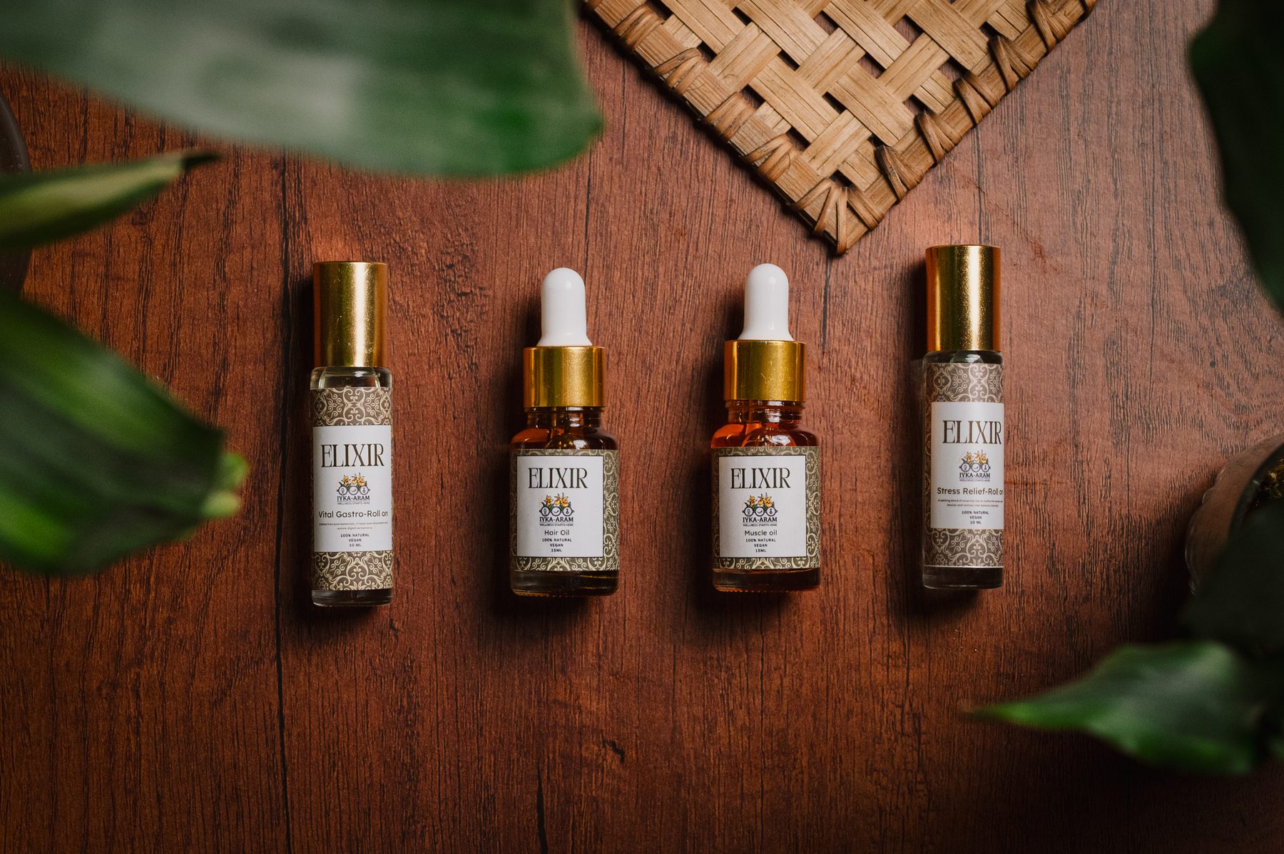 Four ELIXIR bottles laid out on a dark wood table — Vital Gastro Roll-On, Hair Oil, Muscle Oil and Stress Relief Roll-On — beside a woven bamboo mat and tropical leaves