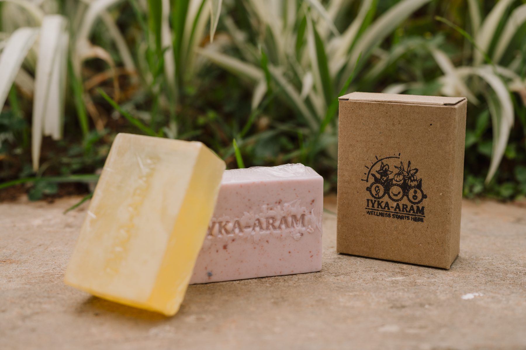 Two Iyka-Aram soap bars — one pale yellow, one rose-flecked pink — each embossed with the brand name, standing beside a stamped kraft carton on a stone ledge