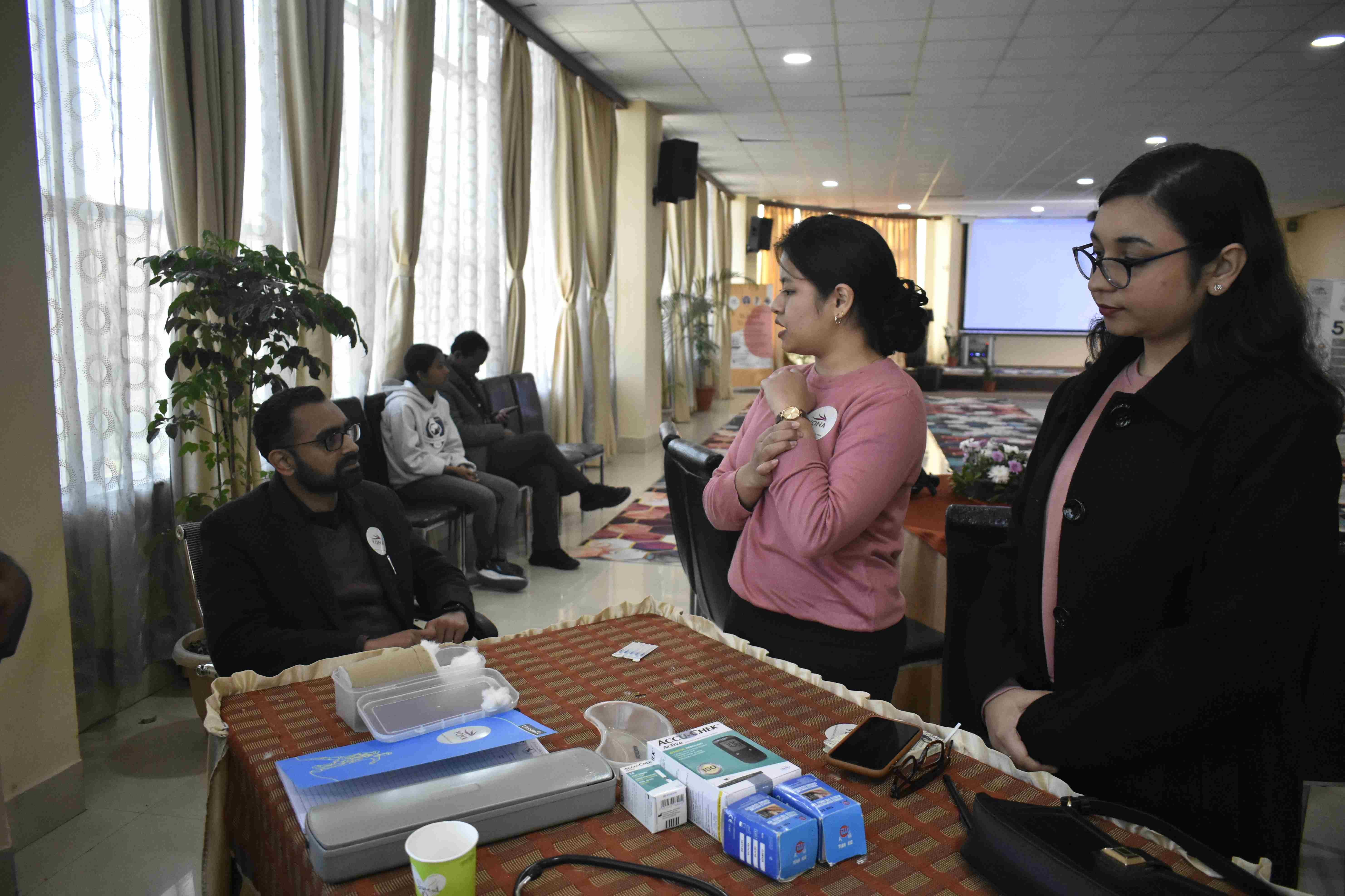 A health screening station at a community wellness camp — a practitioner seated at a table with medical equipment while two staff members assist a participant