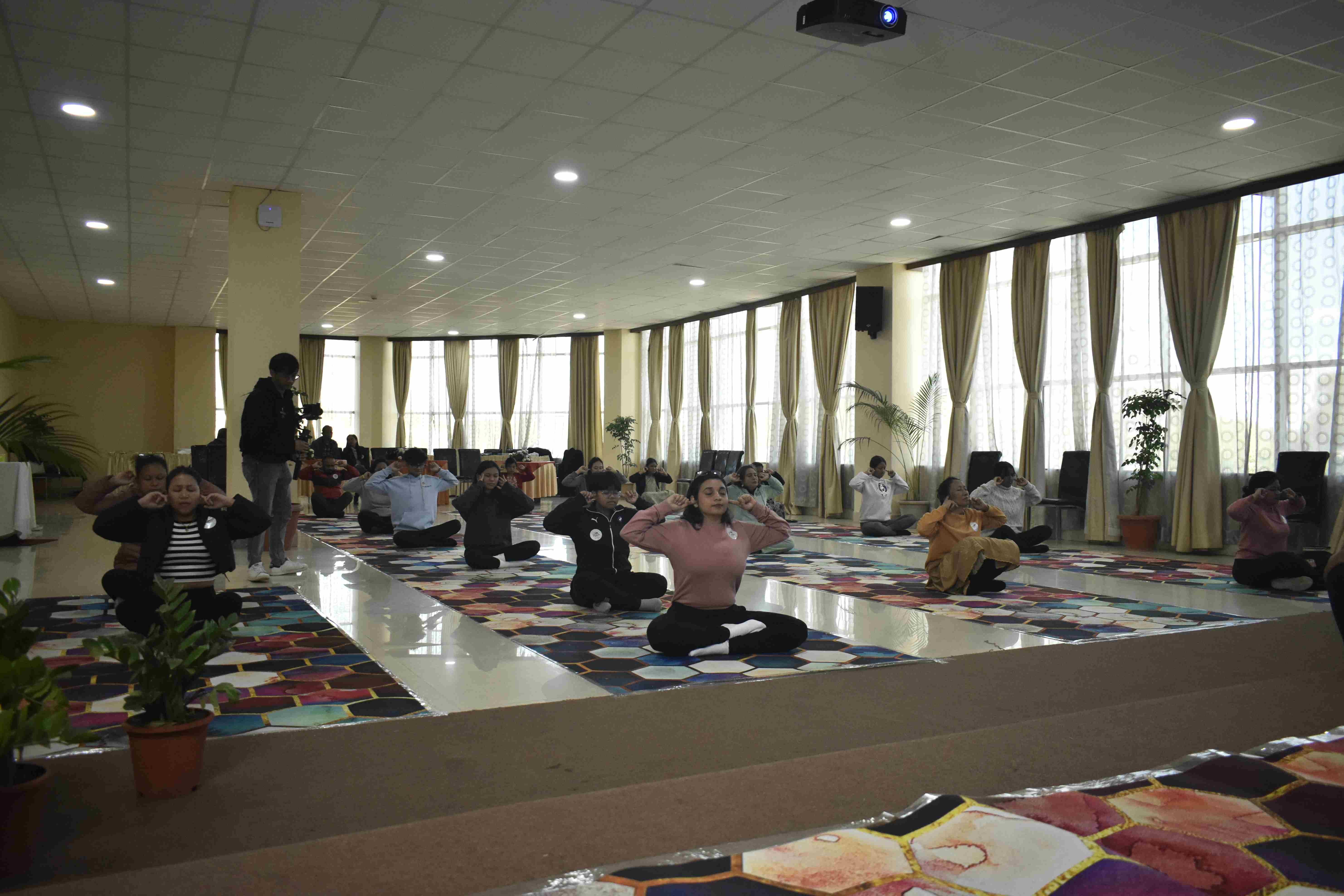 Participants seated in a meditative posture on colourful yoga mats during a large group yoga session in a bright hall with floor-to-ceiling windows