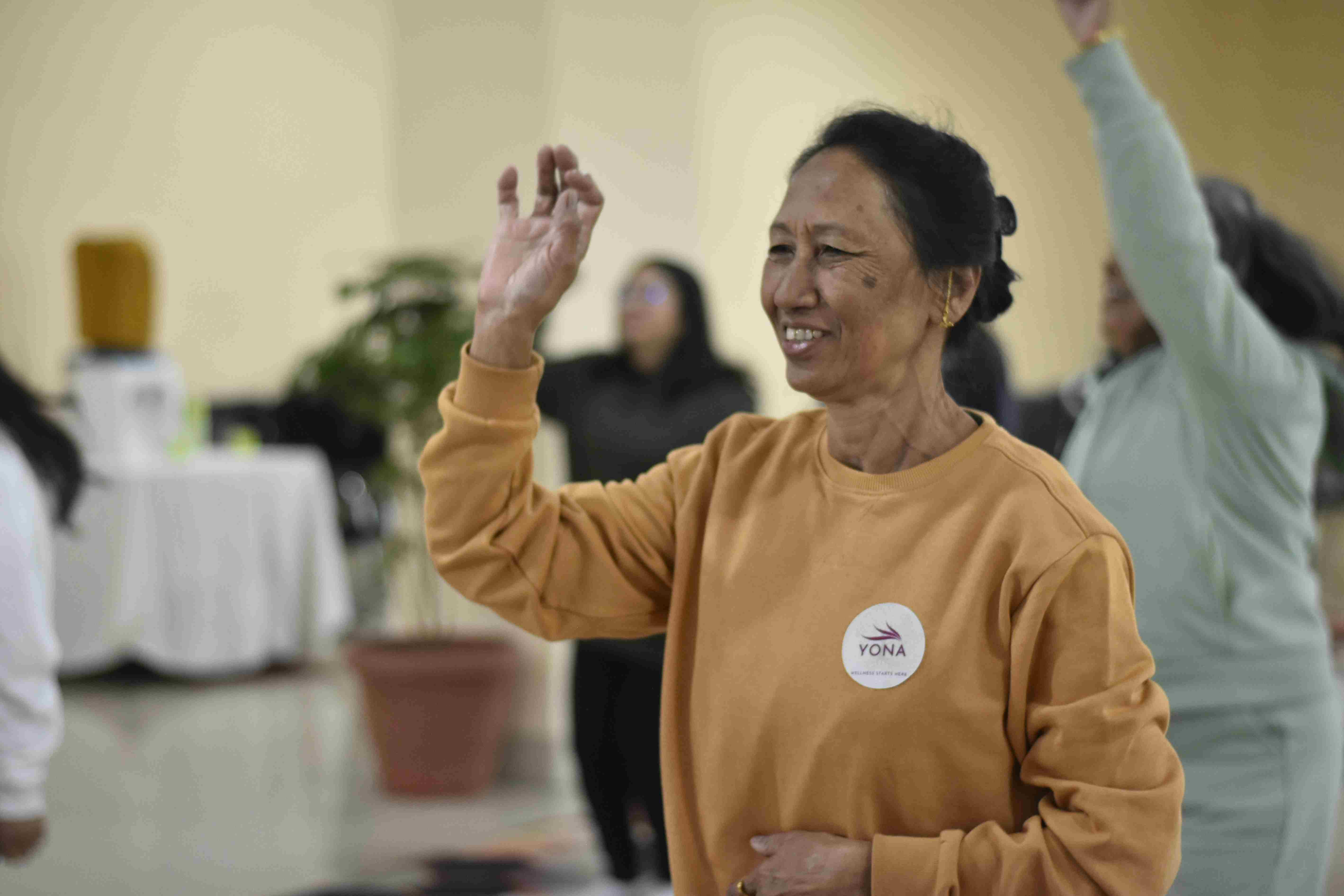 A smiling woman in a mustard-yellow YONA sweatshirt holding a mudra gesture mid-movement during a yoga wellness session