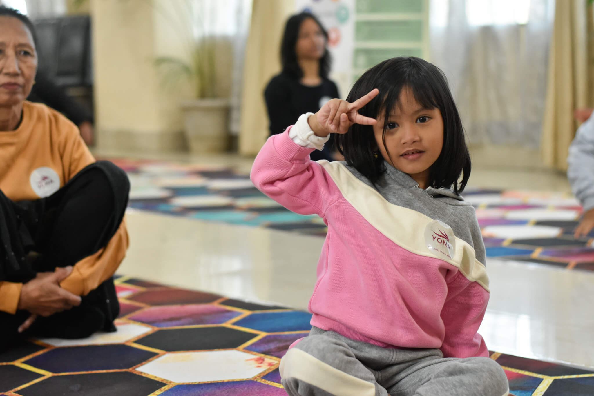 A young girl in a pink YONA sweatshirt sitting cross-legged on a colourful yoga mat making a peace sign during a YONA wellness session