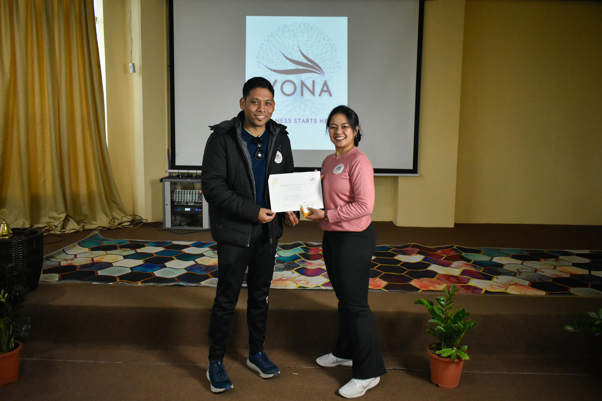 A participant receiving a YONA Certificate of Participation from a female instructor in front of a projected YONA logo at a wellness event