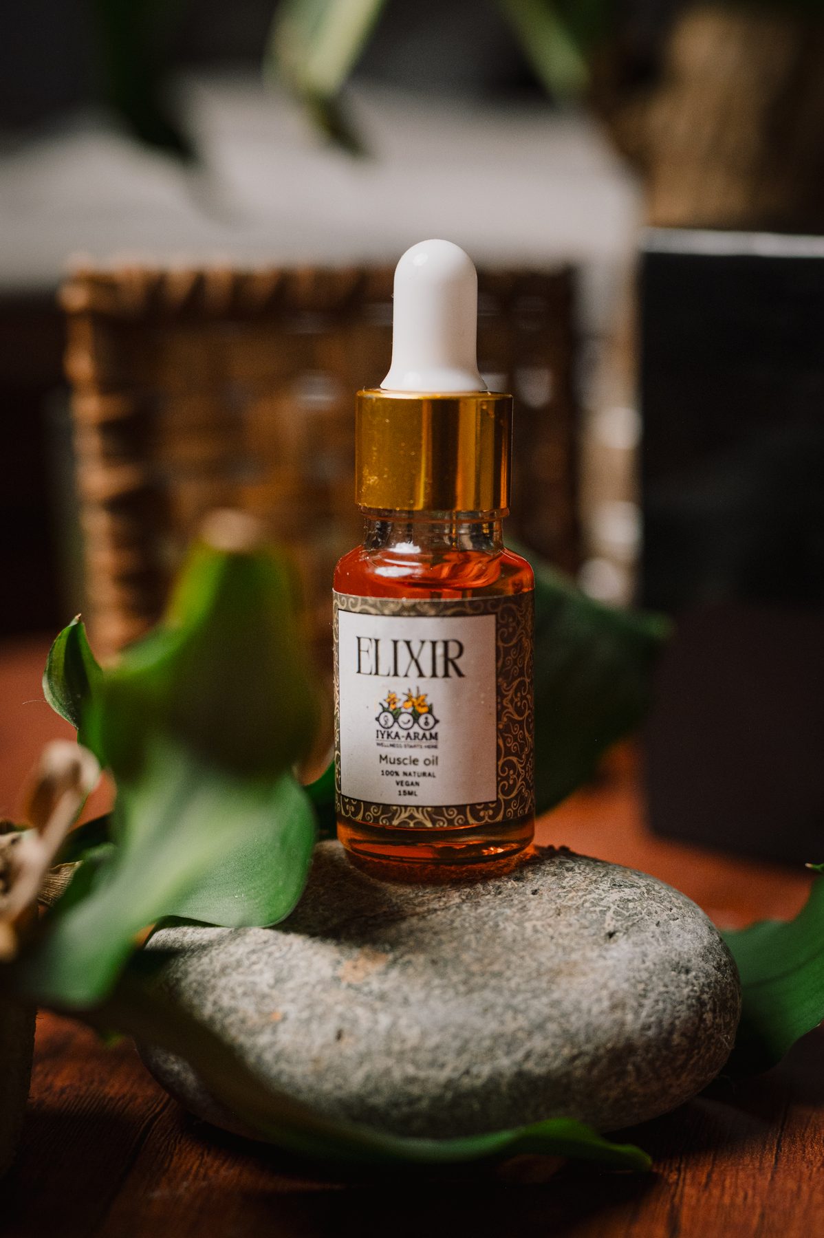 A 15 ml bottle of ELIXIR Muscle Oil with a gold-collared dropper cap resting on a smooth river stone, lit warmly against a dark wooden table