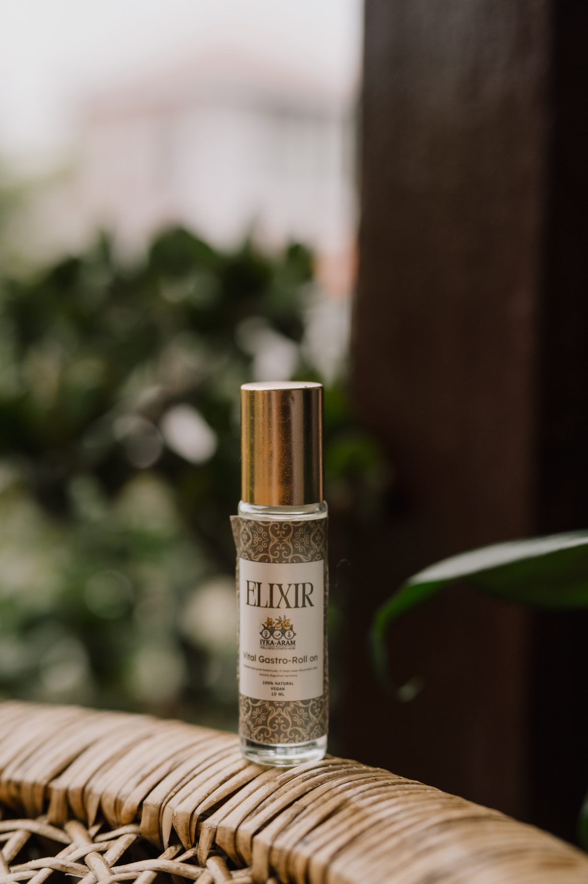 A single ELIXIR roll-on bottle with a gold cap standing on the arm of a woven rattan chair, framed by soft green foliage