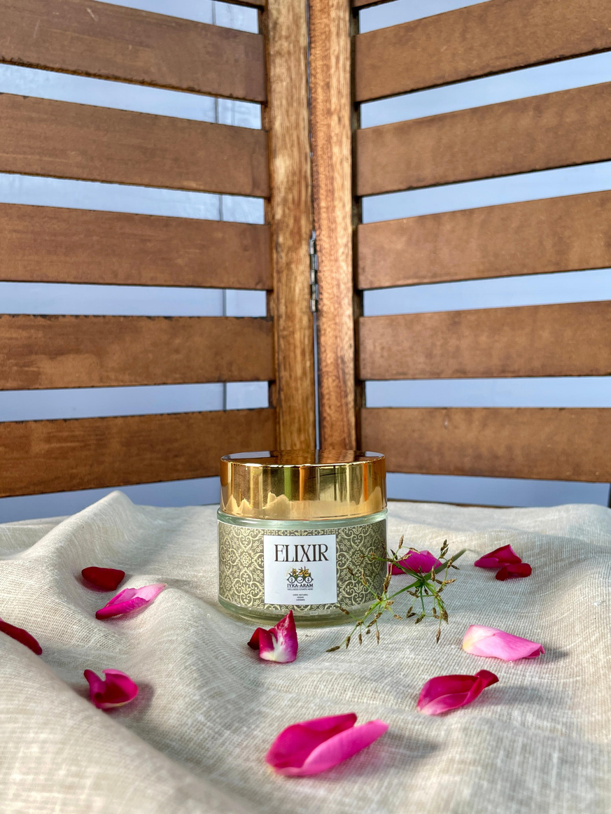 A gold-lidded ELIXIR Aura Cleansing Bath Salt jar standing on cream linen scattered with pink rose petals, photographed against a wooden screen
