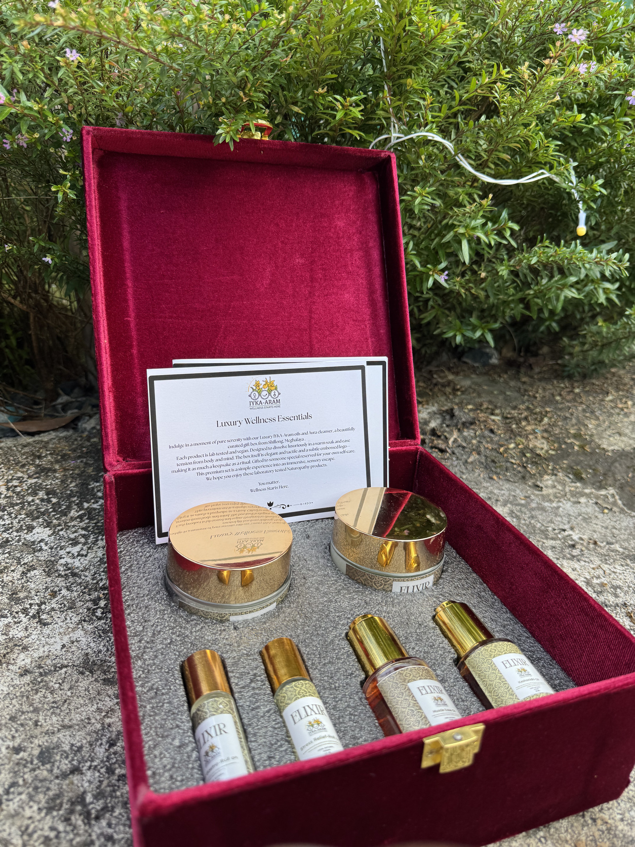 An open crimson velvet Iyka-Aram gift box holding four ELIXIR roll-ons, two gold-lidded jars and a printed Luxury Wellness Essentials card, set outdoors against flowering shrubs