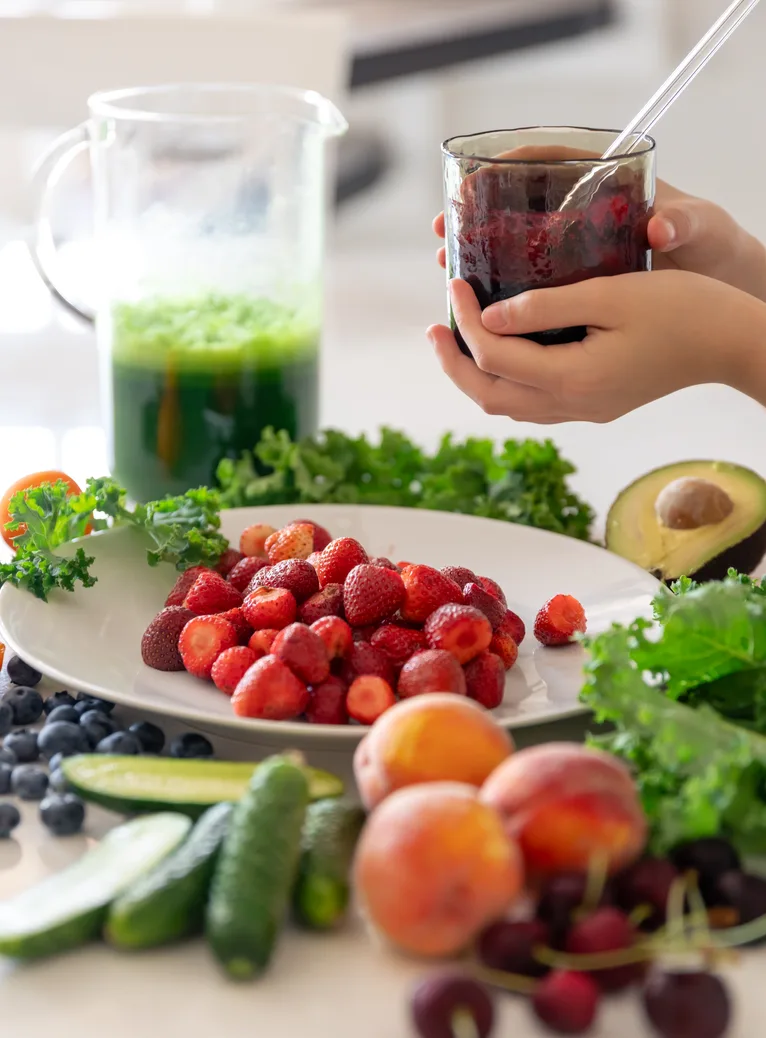 A berry smoothie and green juice beside fresh fruit, greens and avocado on a kitchen counter