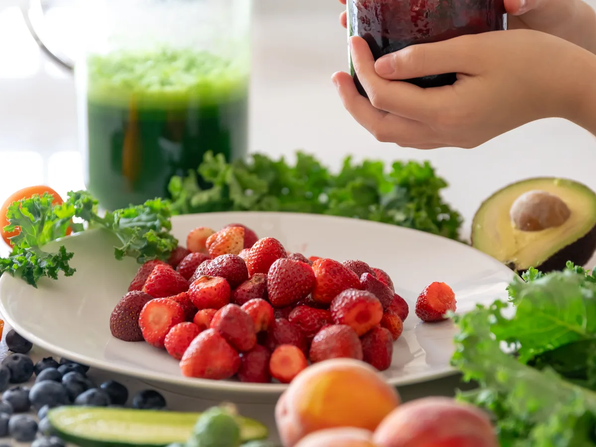A berry smoothie and green juice beside fresh fruit, greens and avocado on a kitchen counter