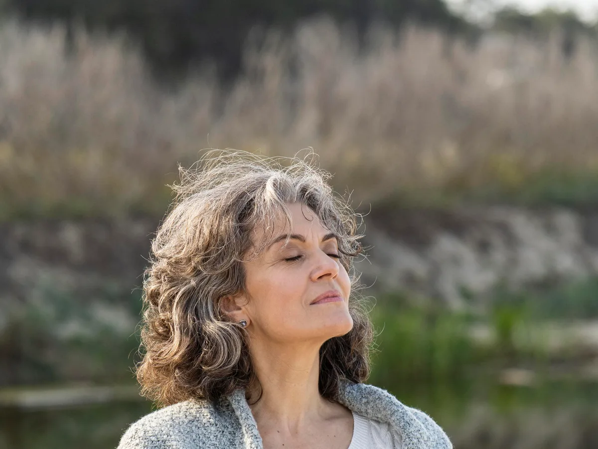 An older woman wrapped in a shawl standing by a lake with her eyes closed